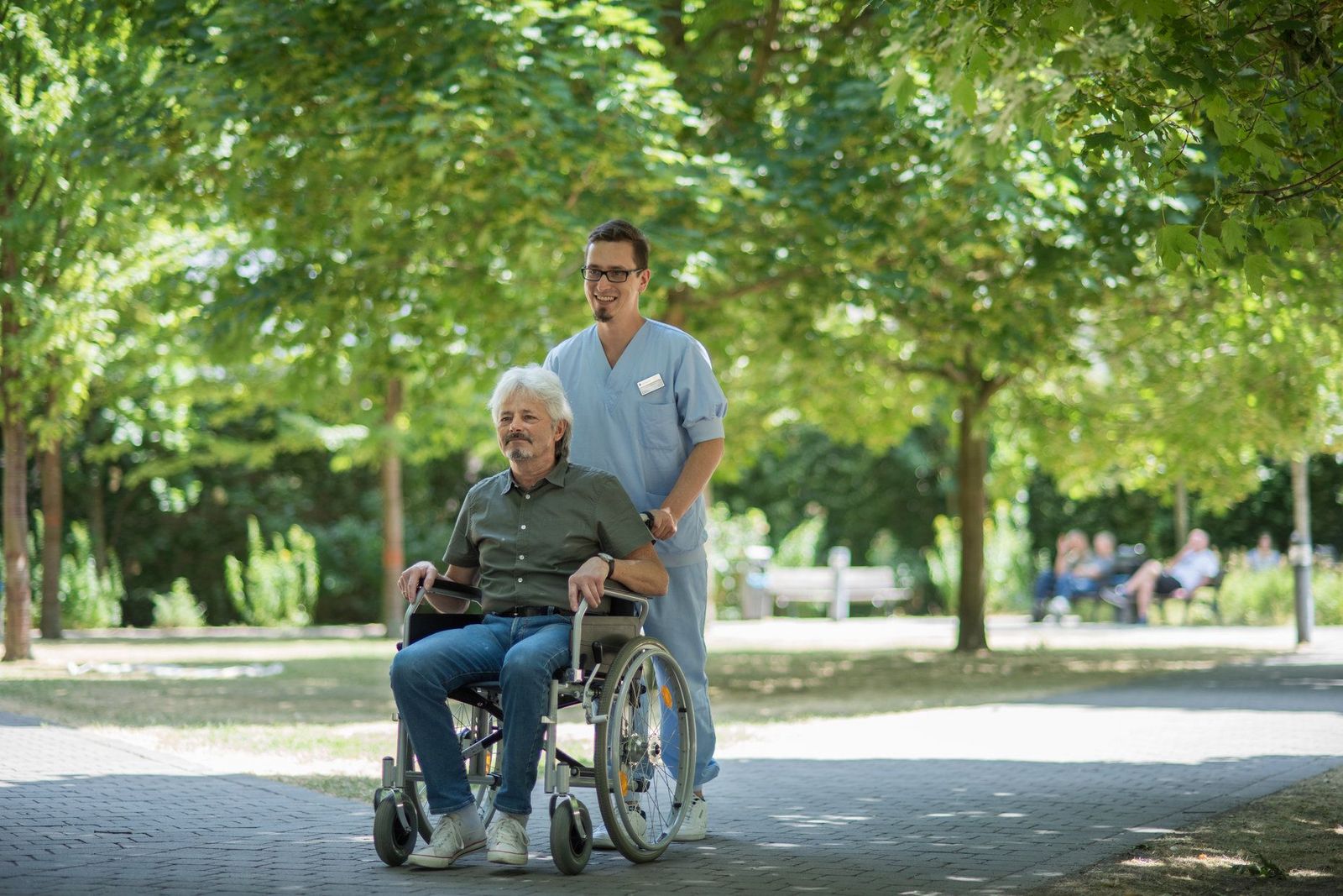 Ein Gesundheits- und Krankenpfleger fährt einen Herrn im Rollstuhl durch den Krankenhausgarten